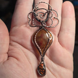 A close-up of a hand holding a copper toned wire-wrapped pendant with a teardrop-shaped amber-brown stone and an ornate spiral wire top.