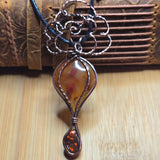 A handmade pendant necklace featuring a teardrop-shaped amber stone with a copper-toned metal frame, suspended from a black cord with a decorative wire top, set against a rustic brown leather and wood background.