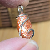 Orange glittery stone pendant with silver wire wrapping held between fingers against a blurred background