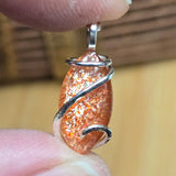 Sparkly orange pendant with silver wire wrapping held between fingers against a blurred background