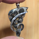 Silver pendant with intricate design held between fingers against a beige background