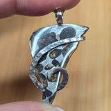 Silver pendant with black speckles held between two fingers against a blurred background
