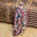 A polished stone pendant with a swirling pattern of red, pink, and white, wrapped in a silver metal band with a gold accent, displayed on a leather and wood background.