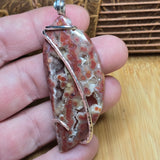 A hand holding a unique polished stone pendant with a silver wire wrap, set against a background of a wooden surface and a woven basket.