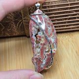 A close-up of a hand holding a carved stone pendant with a silver bail, set against a background of a wooden surface and a leather strap.