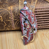 A handcrafted pendant featuring a polished red and white agate slice with organic swirls, wrapped in a dark metal band and a gold-colored curved accent, displayed on a wooden surface with a leather tool bag in the background.