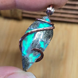 Turquoise Aurora Opal stone pendant with copper wire wrapping held between fingers against a wooden background