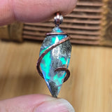 Aurora opal  marcasite agate pendant with copper wire wrapping held between fingers against a wooden background