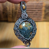 Silver and copper wire-wrapped pendant with a central gemstone held by a hand against a blurred wooden background