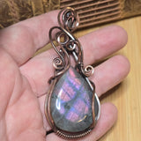 Hand holding a pendant with a rainbow-colored stone and intricate wirework.
