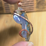 Hand holding a unique pendant with blue stone and copper wire wrapping against a blurred background