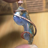 Jewelry pendant with blue stone and copper wire wrapping held in hand against a blurred background