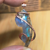Close-up of a pendant with a blue stone and copper wire wrapping, held between fingers.