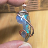 Aurora Opal pendant with copper wire wrapping held between fingers against a wooden background