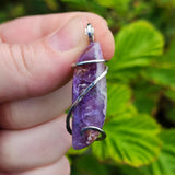Purple amethyst pendant with silver wire wrapping held by a hand against a green leafy background