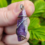 Hand holding a purple gemstone pendant with silver wire wrapping against a green leafy background