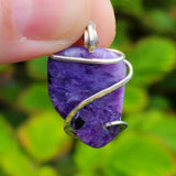 Purple gemstone pendant with silver wire wrapping held by a hand against a green leafy background