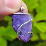 Purple stone pendant with silver wire wrapping held by a hand against a green leafy background