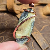 Jewelry with a stone and copper wire wrapping held by a hand on a textured surface.