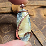 Plume Agate pendant with copper wire wrapping held between fingers against a textured brown background