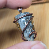 Pendant with stone and copper wire wrapping held between fingers against a wooden background