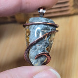 Close-up of a stone pendant with copper wire wrapping held between fingers on a wooden surface.