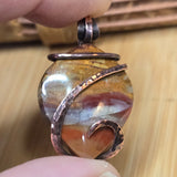 Close-up of a copper-wrapped stone pendant held between fingers on a wooden surface.
