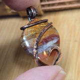 Close-up of a stone pendant with copper wire wrapping held by a hand on a wooden surface.