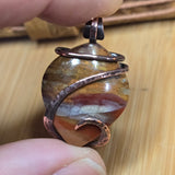 Close-up of a stone pendant with copper wire wrapping held by a hand on a wooden surface.