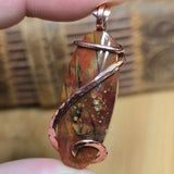 Hand holding a copper-wrapped pendant with a natural stone design against a blurred background
