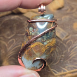 Pendant with natural stone and copper wire wrapping held by a hand against a textured background