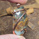 Jasper pendant with copper wire wrapping held by a hand against a textured brown background