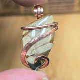 Pendant with stone and copper wire wrapping held between fingers against a blurred background
