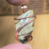 Pendant with stone and copper wire wrapping held between fingers against a blurred background