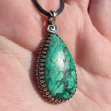Teardrop-shaped green stone pendant on a black cord held in hand