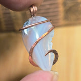 Pendant with a white stone and copper wire wrapping held between fingers against a blurred background