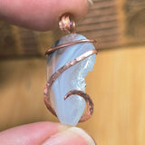 Pendant with a light purple stone and copper wire wrapping held between fingers against a blurred background