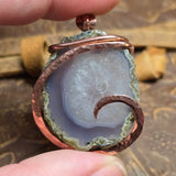 Pendant with agate stone and copper wire wrapping held by a hand against a textured background