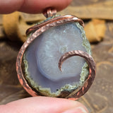 Close-up of a copper-wrapped agate pendant held between fingers with a natural stone background.