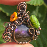 Decorative pendant with purple stone, orange flower, and green leaf against a blurred green background