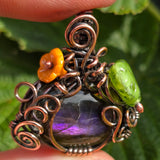 Ornate wire-wrapped pendant with a central purple stone, orange and green beads, held against a green leafy background.
