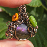 Ornate pendant with a purple stone, orange flower, and green insect held between fingers against a leafy background