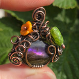 Ornate copper pendant with a purple gemstone, orange flower, and green leaf held between fingers against a green leafy background.