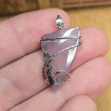 A hand holding a triangular lavender-purple stone pendant wrapped with silver wire, shown close-up against a warm wooden background.