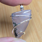 A close-up of a hand holding a triangular polished stone pendant wrapped in silver wire, set against a light wood background.