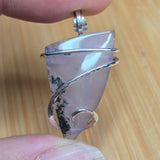 A close-up of a hand holding a silver-wrapped pendant featuring a translucent lavender-gray stone with dark inclusions, set against a light wood background.