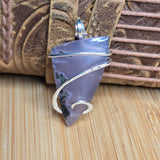 A handmade silver wire-wrapped pendant featuring a polished, irregular-shaped lavender-purple stone with natural green inclusions, displayed on a wooden surface with a brown leather strap in the background.