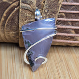 A handmade silver wire-wrapped pendant featuring a polished, irregular-shaped lavender-gray stone with natural dark inclusions, resting on a light wood surface in front of a brown leather strap.