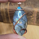 Labradorite pendant with copper wire wrapping held between fingers against a blurred background