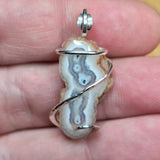 A close-up of a hand holding a silver-wrapped agate stone pendant. The pendant features natural banded agate patterns in white, gray, and brown, with a polished silver bail at the top.